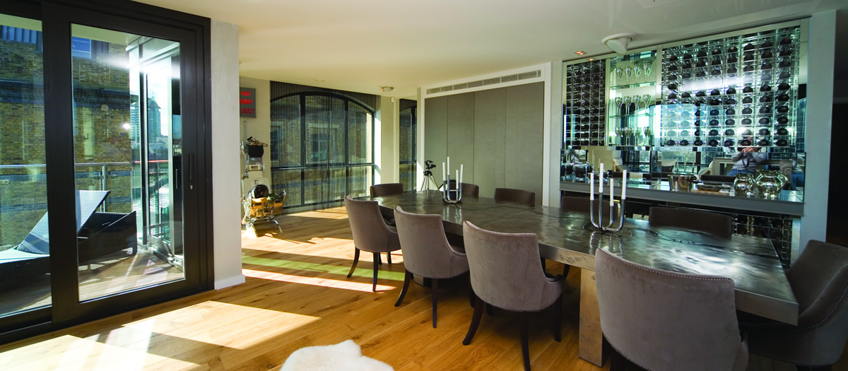 Bright entertaining/dining space with glass doors to a balcony, a long table with upholstered chairs, and a mirrored wine wall and bar.